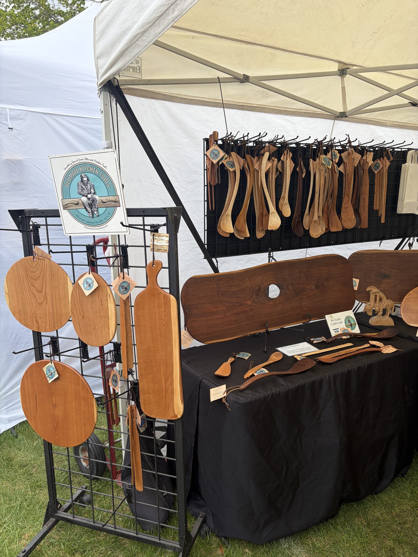 The Ultimate Kitchen Tools booth at an art show, with carved spoons hanging on a rack and serving boards on display