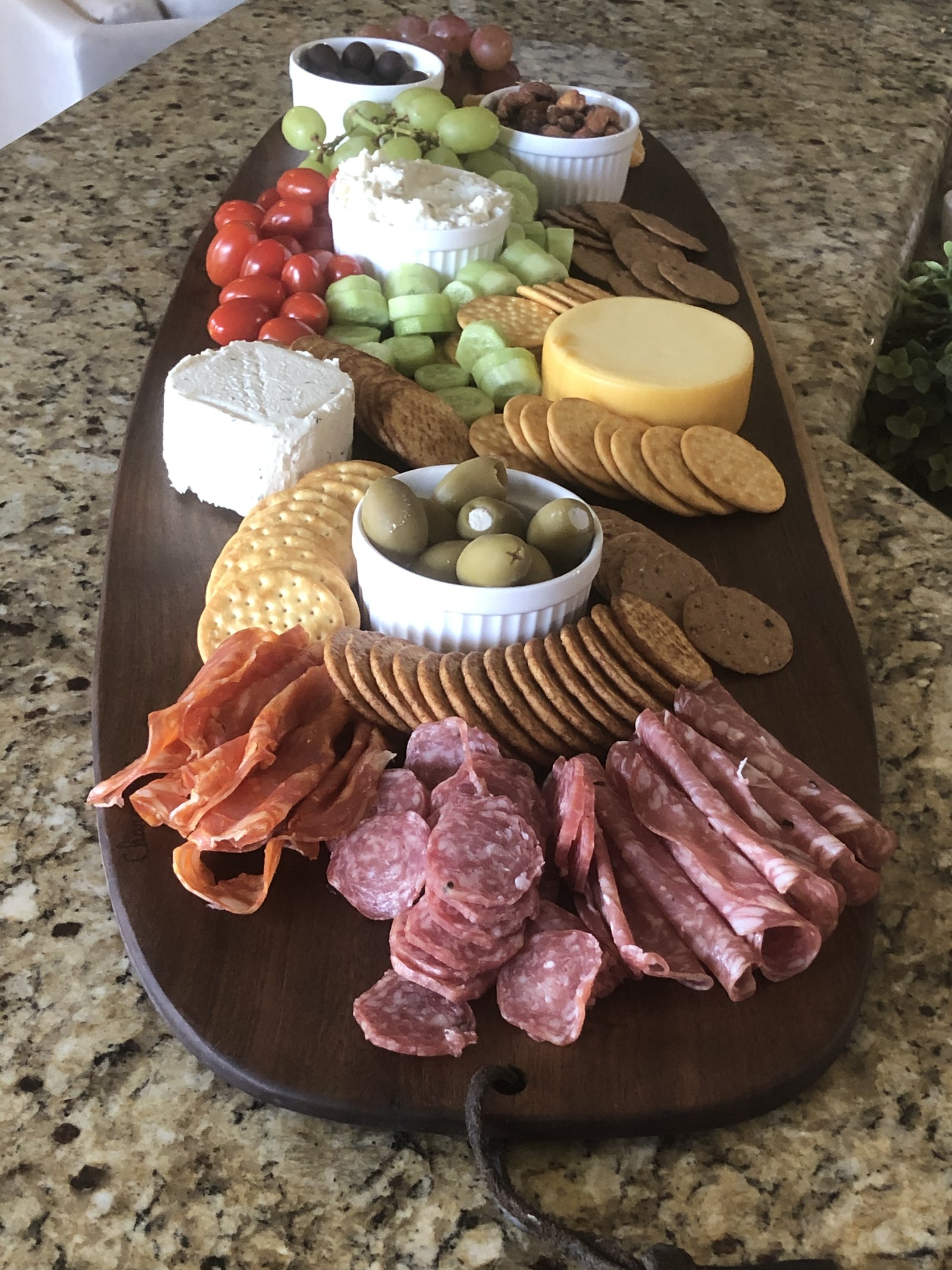 A long walnut charcuterie board on a kitchen counter, loaded with sliced salami and prosciutto, crackers, cheeses, olives, grapes, tomatoes and cucumber