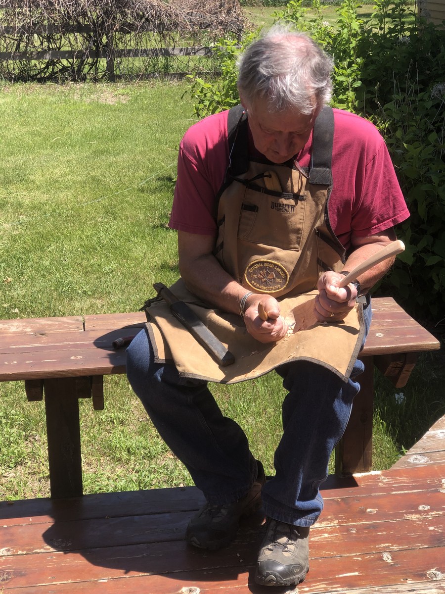 Chris Rounds sitting on a bench outdoors in his work apron, shaping a wooden spoon with a carving knife, shavings scattered on the deck