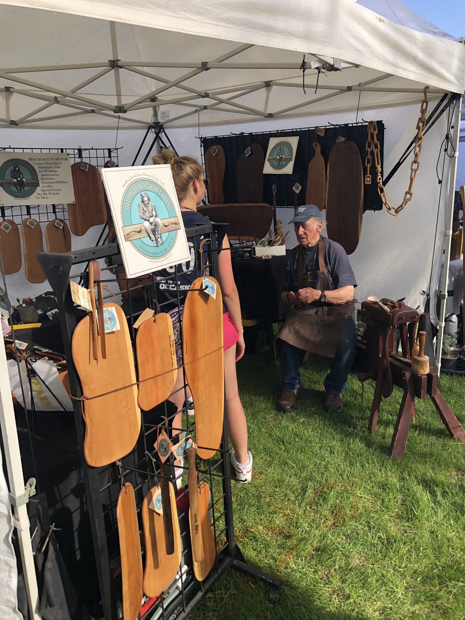 Chris Rounds in a leather apron, carving at his booth beside a shaving horse, with boards and tools on display