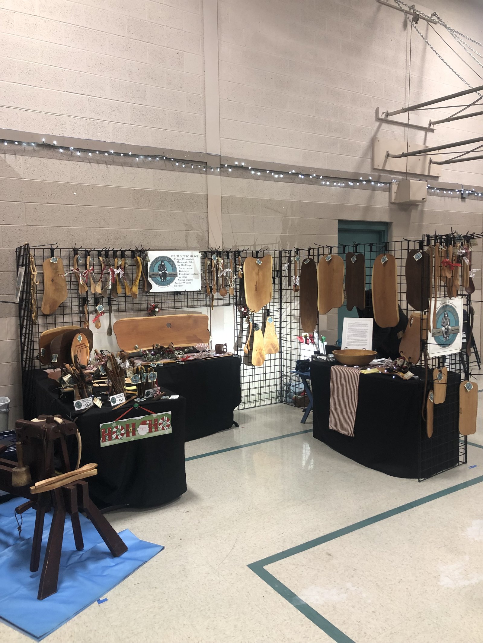 The full Ultimate Kitchen Tools booth at a holiday show, two tables of carved tools under grid walls hung with serving boards, with the shaving horse set up in front