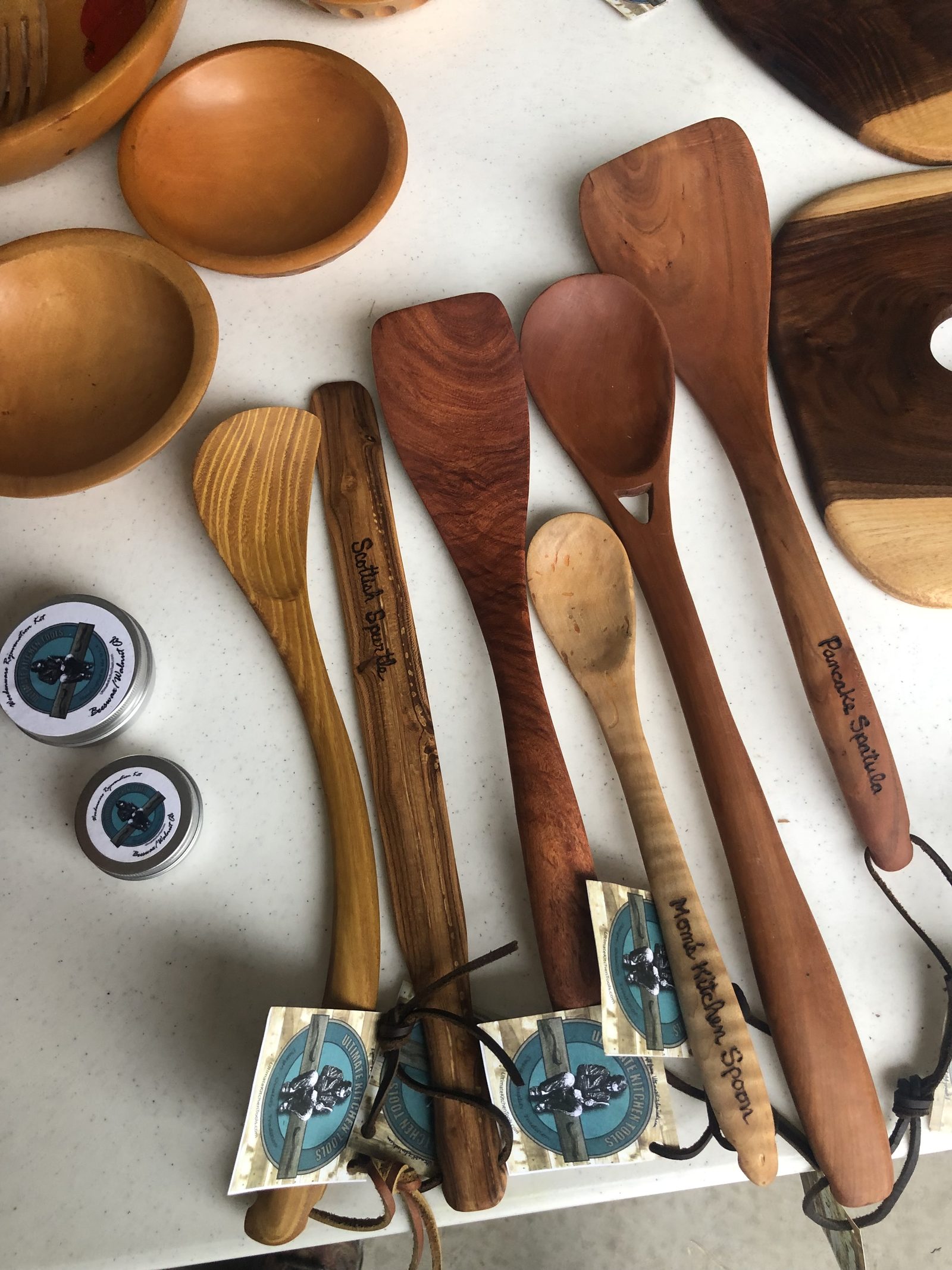 Carved spoons, spatulas and a spurtle laid out beside wooden bowls and maintenance kit tins, several with their names burned into the handle