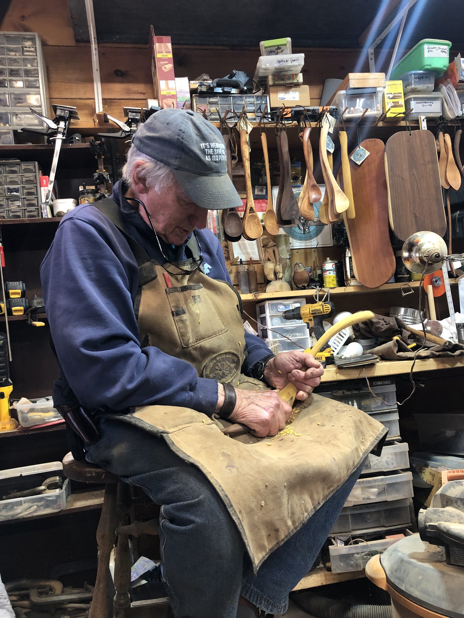 Chris Rounds carving a wooden spoon in his workshop, with finished spoons and boards hanging on the wall behind him
